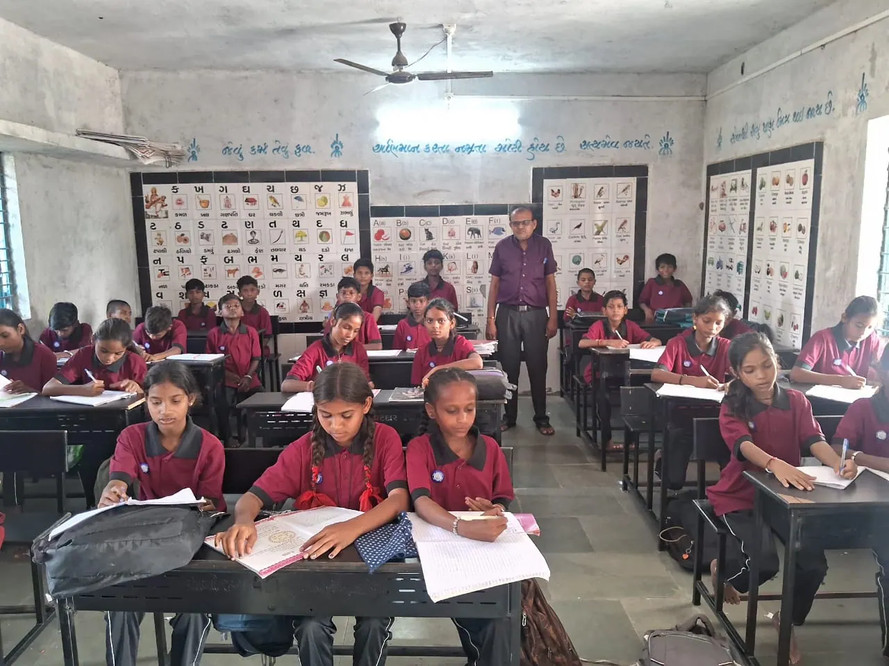 Students writing at their desks while a teacher stands between the rows, with alphabet charts on the classroom wall.