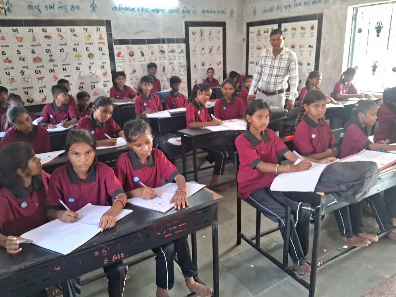 Girls in school uniform writing in their notebooks during a lesson as a teacher looks on.