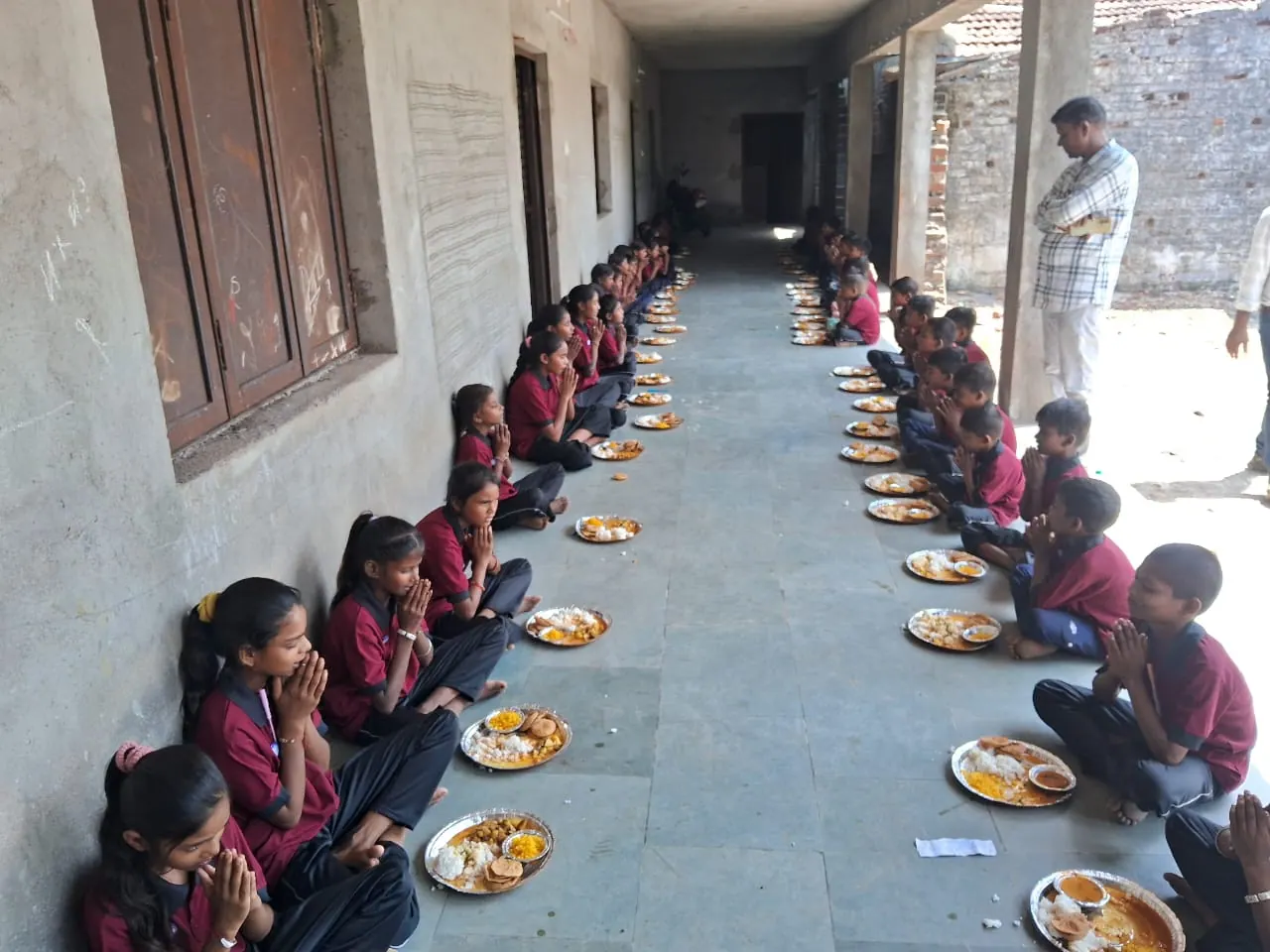 Students seated in a row along the school verandah, hands folded in prayer before a meal.