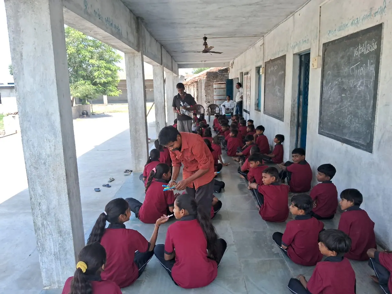 A staff member handing out pens to students seated on the school verandah.