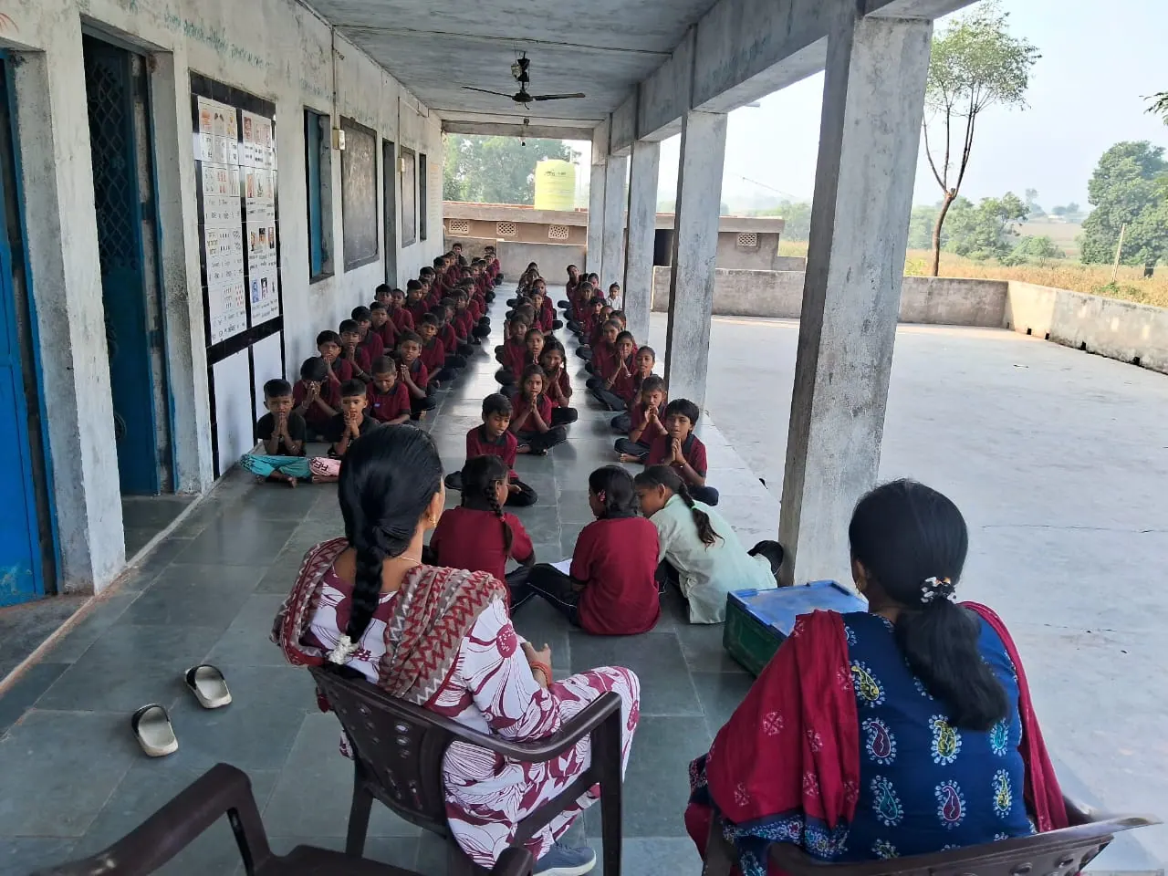 Two teachers seated on the verandah watching over students gathered in rows.